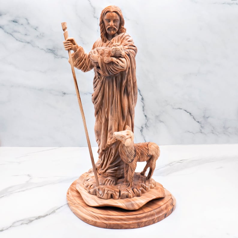 Op de afbeelding: Een houten beeldhouwwerk dat een figuur voorstelt die een lam en een staf vasthoudt, met een ander lam aan zijn voeten. Het beeld is gemaakt van lichtbruin hout en staat op een ronde basis. De figuur heeft lang haar en een baard.