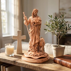 Op de afbeelding: Een houten beeld van een religieuze figuur met een staf en een lam, met een kruis en een brandende kaars op een houten tafel. Een olijfboom in een pot en een boek staan ook op de tafel. Het beeld is lichtbruin.