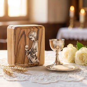 May include: A wooden box with a silver relief of a kneeling child holding a chalice and grapes. A silver chalice, paten with a host, and wheat stalks are on a white lace tablecloth. Two white roses and lit candles are in the background.