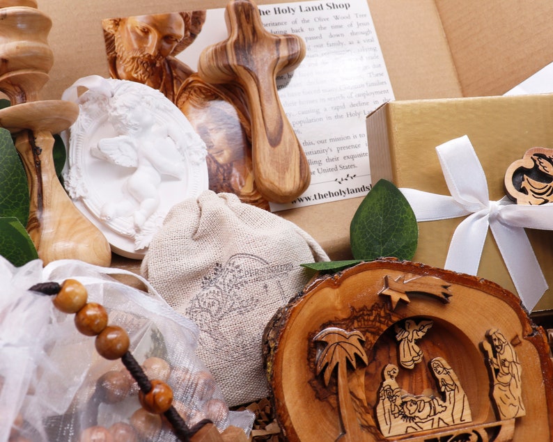 May include: An assortment of religious items, including a wooden cross, a carved nativity scene, and a white angel relief. A small burlap bag and a gold gift box with a white ribbon are also visible. The items are displayed in a cardboard box.