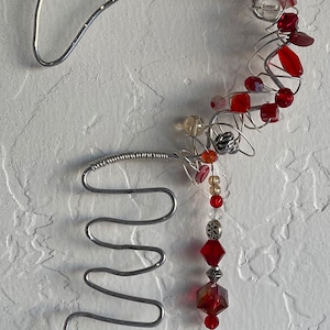 May include: A silver wire heart-shaped ornament with red and clear glass beads. The ornament has a spiral design at the bottom and two dangling heart-shaped beads.