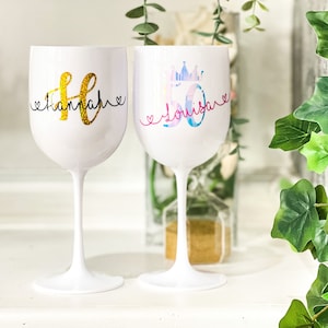 May include: Two white wine glasses with personalised designs. The first glass has a gold glitter design with the name "Hannah" and a heart on each side. The second glass has a pink and blue design with a crown and the number "50" and the name "Louise" with a heart on each side.