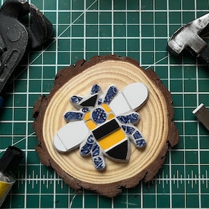 May include: A mosaic bee figure crafted from blue and white patterned ceramic pieces, solid yellow, and black tiles. The bee is centred on a circular natural wood slice with visible growth rings, surrounded by metal pliers, a hammer, and glue.