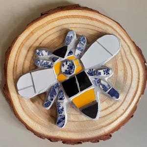 Puede incluir: Una abeja de mosaico hecha de azulejos de cerámica azul y blanco, con un abdomen rayado amarillo y negro, está montada en una rodaja de madera.