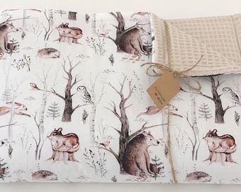 Op de afbeelding: Een witte babydeken met een print van bosdieren. De deken heeft beren, herten, uilen en egels in aquarelstijl. De deken is vastgebonden met een bruin lint en een label met de tekst "Handmade with Love".