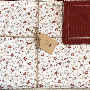 Könnte beinhalten: Ein Set aus zwei weißen Stoff-Geschirrtüchern mit einem kleinen Blumenmuster in Rot-, Rosa- und Gelbtönen. Die Tücher sind mit Schnur zusammengebunden und mit einem braunen Etikett versehen, auf dem "mit Liebe gemacht" steht. Ein rotes Waffelgewebe-Geschirrtuch ist im Set enthalten.