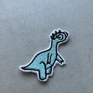 May include: A light blue embroidered iron-on patch of a cartoon dinosaur with white outlines. The dinosaur has a large eye and a spiky crest.