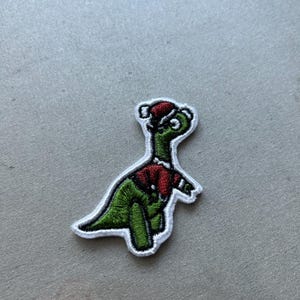 May include: A green dinosaur wearing a red Santa hat and a red and white sweater. The dinosaur is embroidered on a white fabric patch.