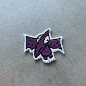 May include: Purple embroidered iron-on patch of a cartoon bat with white outlines.