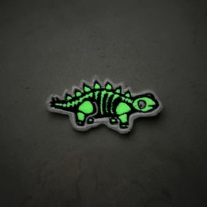 May include: A green glow-in-the-dark dinosaur skeleton patch. The patch is embroidered with white thread and has a white border.