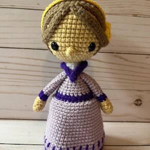 May include: A crocheted doll with brown hair, a yellow headband, a purple dress, and black eyes.