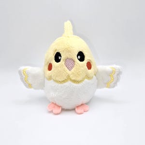 May include: A plush toy cockatiel in a cheerful design. The bird features a yellow crest and head, black eyes, and orange cheek spots. Its wings are white with yellow trim, and it has pink feet. The toy is soft and cuddly, perfect for children.