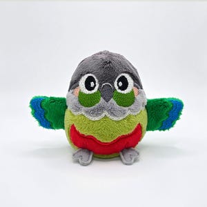 May include: A plush bird toy featuring a gray head, green cheeks, and a lime green body with a red belly patch. The wings are green with blue tips, and the bird has large white eyes with black pupils and small gray feet extending from the base.