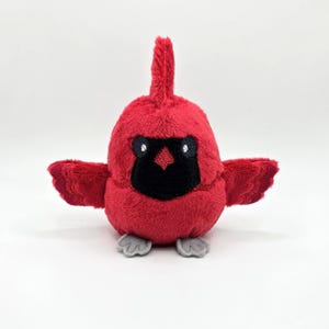 May include: A plush red cardinal bird with a black face and small gray feet. The bird has small wings and a crest on its head. The plush toy is made of soft material and is designed to look like a cardinal.