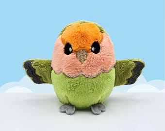 May include: A plush bird toy featuring a bright orange forehead, a soft pink face, and a light green body. The bird has small black embroidered eyes, a tan beak, and outstretched wings with dark gray tips. It stands on two small gray felt feet.