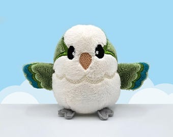 May include: A small plush bird toy with a white body, a tan beak, and black embroidered eyes. The wings are green with blue and dark green accents. The bird has small gray feet and is depicted with its wings spread out to the sides.
