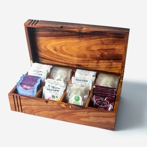 May include: A wooden tea box with a hinged lid, open to reveal compartments filled with various tea bags. The tea bags are labelled with brand names such as "Yogi", "Bigelow", and "Private Selection".