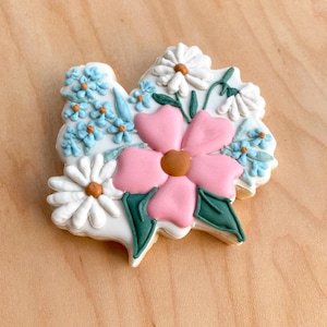 Peut inclure: Un biscuit décoré avec une fleur rose, des marguerites blanches et des fleurs bleues. Le biscuit est décoré de feuilles vertes et de centres marrons.