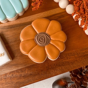 May include: A flower-shaped biscuit with orange icing petals and a brown centre. The biscuit sits on a wooden board, alongside other autumnal decorations.