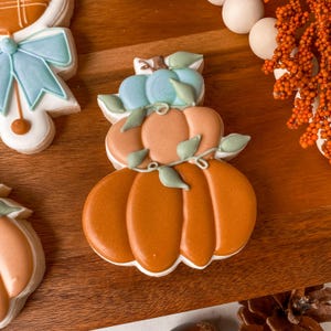 May include: A stack of three pumpkin-shaped cookies with orange icing, green leaf accents, and a light blue pumpkin on top. The cookies are decorated with white icing outlines and sit on a wooden surface.
