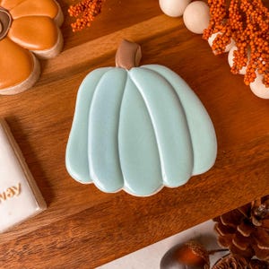 May include: A light blue pumpkin-shaped cookie with a brown stem. The cookie is decorated with icing and sits on a wooden surface, alongside other fall-themed cookies and decorations. The overall theme is autumn and fall treats.
