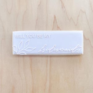 May include: White rectangular cookie with the text "Will you be my bridesmaid?" and a simple line drawing of a leaf branch.