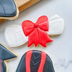 May include: Graduation-themed cookies. A white diploma cookie with a red bow is the focal point. Other cookies include a black graduation cap and a mortarboard. The cookies are decorated with white and red icing.