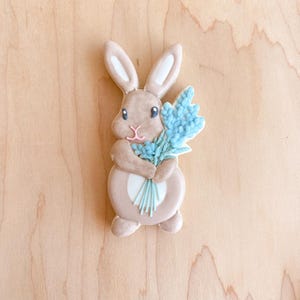 Puede incluir: Una galleta de conejo marrón sosteniendo un ramo de flores azul claro. La galleta está decorada con glaseado y tiene una mancha blanca en el vientre.