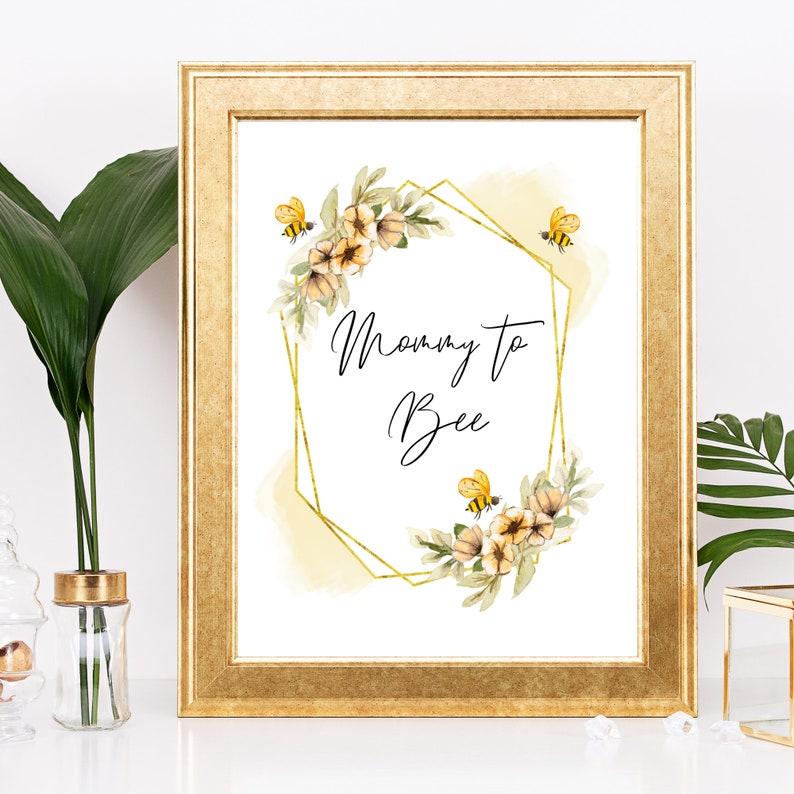 May include: A gold frame with a white background and a watercolor illustration of yellow flowers and bees. The text "Mommy to Bee" is written in black cursive font.