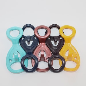 May include: Five colorful plastic carabiner-style clips in shades of teal, navy, rose, dark brown, and yellow. Each clip has a rounded top loop and a lower oval loop, with a central design. These clips are likely for outdoor or sporting use.