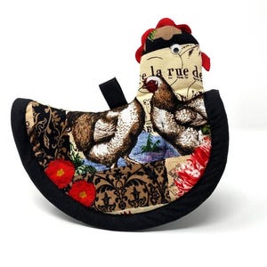 May include: A black and white patterned fabric hot pad shaped like a chicken with a red comb and a large black eye. The fabric features a French phrase "e la rue de" and images of chickens.