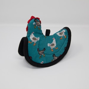 Puede incluir: Un agarrador de olla de color azul turquesa en forma de pollo con ribete negro. El agarrador de olla está decorado con un patrón de pollos blancos y negros.