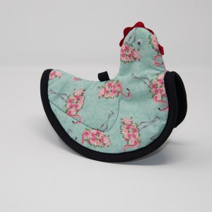 May include: A teal blue hot pad shaped like a chicken with a red comb and black trim. The hot pad is decorated with a pattern of pink flamingos with floral accents.