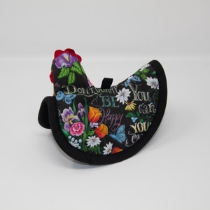 May include: A black chicken-shaped oven mitt with a floral and butterfly pattern. The mitt has a red comb and a black loop for hanging. Text on the mitt reads "Don't worry, be happy. You can, you will."