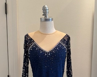 May include: A navy blue figure skating dress with long sleeves and a V-neckline. The dress is embellished with rhinestones and features a lace overlay on the sleeves. The dress has a fitted bodice and a flared skirt.