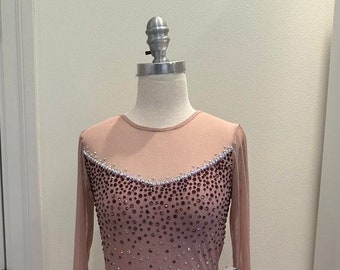 May include: A pale pink figure skating dress with long sleeves and a beaded bodice. The dress has a sheer overlay and a small tag that reads "Winter Sparkle Studios".