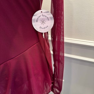 May include: A burgundy long-sleeved mesh leotard with a ruffled hem. The leotard has a white tag attached with the text "Winter Sparkle Studios".