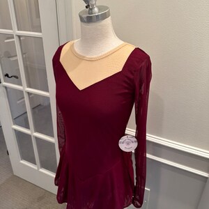 May include: Burgundy figure skating dress with long sleeves and a sheer beige mesh panel at the neckline. The dress has a fitted bodice and a flared skirt. The dress is on a white mannequin.