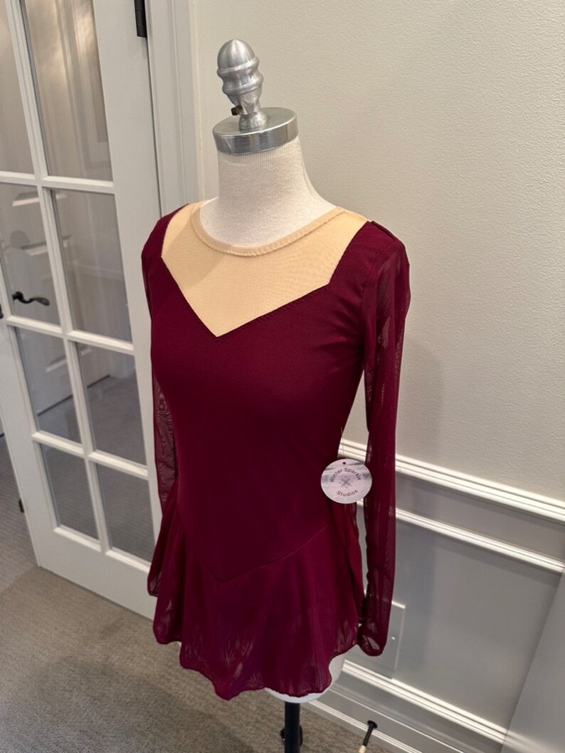 May include: Burgundy figure skating dress with long sleeves and a sheer beige mesh panel at the neckline. The dress has a fitted bodice and a flared skirt. The dress is on a white mannequin.