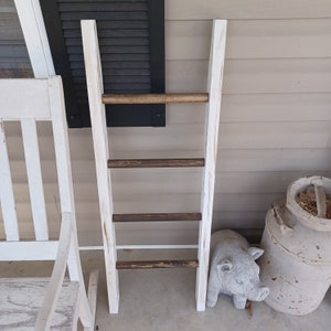 May include: White wooden ladder shelf with four brown wooden rungs. The ladder shelf is standing on a white wooden floor.