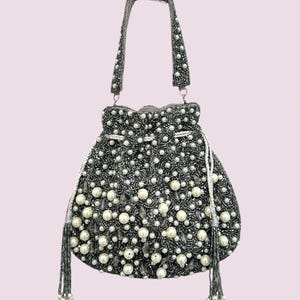 May include: A drawstring handbag featuring a dark gray beaded exterior embellished with numerous white faux pearls and clear crystal accents. The bag includes a single beaded handle and two silver-toned drawstrings finished with beaded tassels.