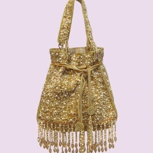 May include: A gold-colored drawstring handbag featuring intricate metallic embroidery, scattered white faux pearls, and a single top handle. The base of the bag is adorned with a fringe of hanging gold beads and clear teardrop-shaped crystals.