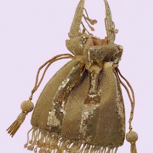 May include: A gold drawstring pouch bag covered in shimmering sequins and beads. The bag features a single top handle, a gathered top closure with two long braided cords ending in beaded tassels, and a bottom fringe of dangling gold beads.