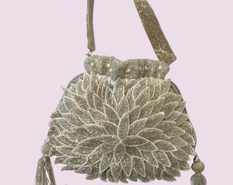 May include: A champagne-coloured beaded handbag featuring a layered petal design on the front. The bag has a single top handle, a drawstring closure with small white pearls, and two decorative tassels with silver-toned accents on the sides.