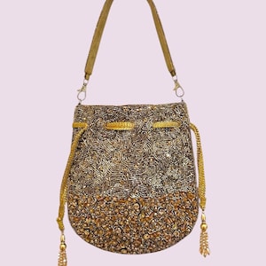 May include: A drawstring handbag featuring a metallic gold and bronze beaded exterior with a swirling pattern on the upper section and larger faceted amber-toned beads on the lower portion. It includes a gold shoulder strap and two side tassels.