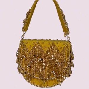 Può includere: Una borsa a mano color giallo senape con silhouette semicircolare, decorata con perline di cristallo trasparenti e dorate a cascata. Il manico è impreziosito da perline abbinate e la patta frontale presenta un motivo a zigzag di cristalli.