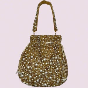 May include: A gold-toned handbag featuring an intricate mesh of gold beads and scattered white faux pearls. The bag has a single top handle adorned with matching beads and pearls, a drawstring closure with gold tassels, and a structured base.