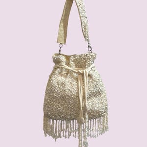 May include: Ivory beaded drawstring bag with a matching shoulder strap. The bag features a fringe of dangling beads along the bottom and a drawstring closure. The bag is a light cream color and has a textured surface.