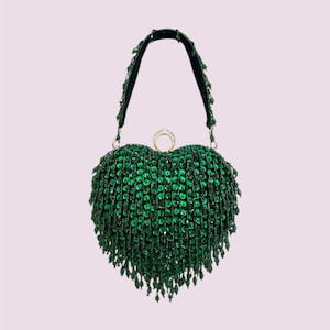 May include: A heart-shaped green beaded handbag with a black handle and gold-colored ring clasp. The bag is covered in small, round, emerald-green beads, with longer beaded fringe at the bottom. The handle is black velvet.
