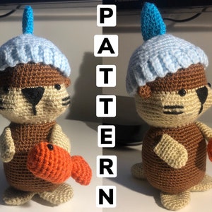 May include: A crocheted otter wearing a blue and turquoise hat and holding an orange crocheted fish. The otter is brown with black eyes and a black nose. The word "PATTERN" is written vertically in black letters on the right side of the image.